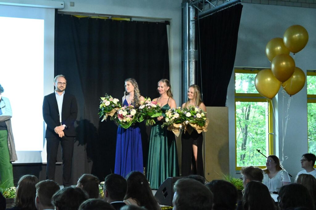 Drei Frauen in eleganten Kleidern halten Blumensträuße auf der Bühne.