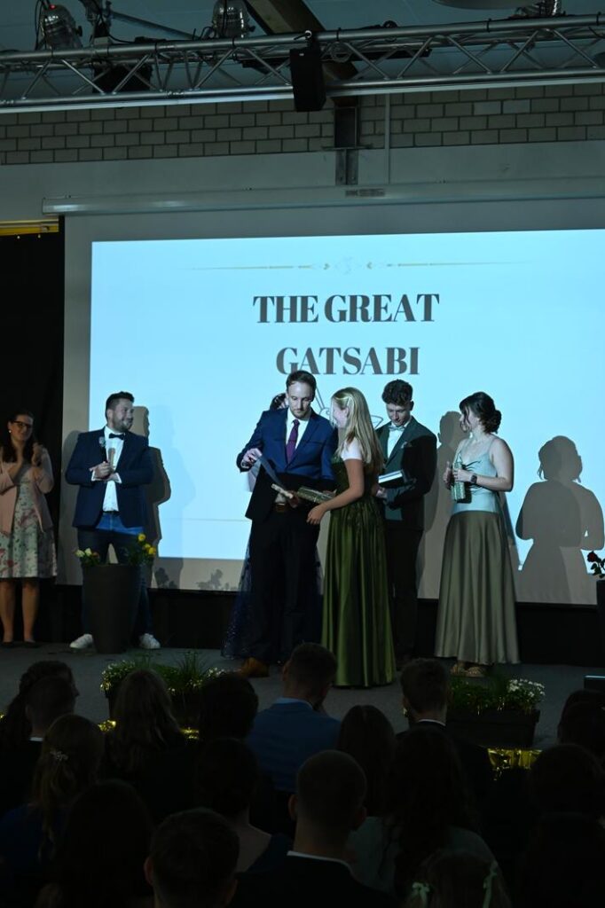 Ein Festakt mit mehreren Personen auf der Bühne vor einem Bildschirm mit "THE GREAT GATSABI".