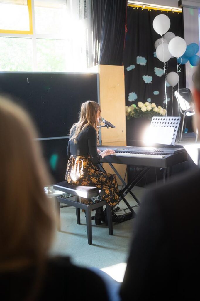 Eine junge Frau spielt Klavier und singt, umgeben von Dekorationen und Publikum.