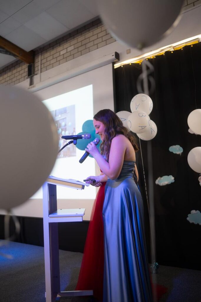 Eine Person in einem eleganten Kleid spricht am Mikrofon vor einer Präsentation.