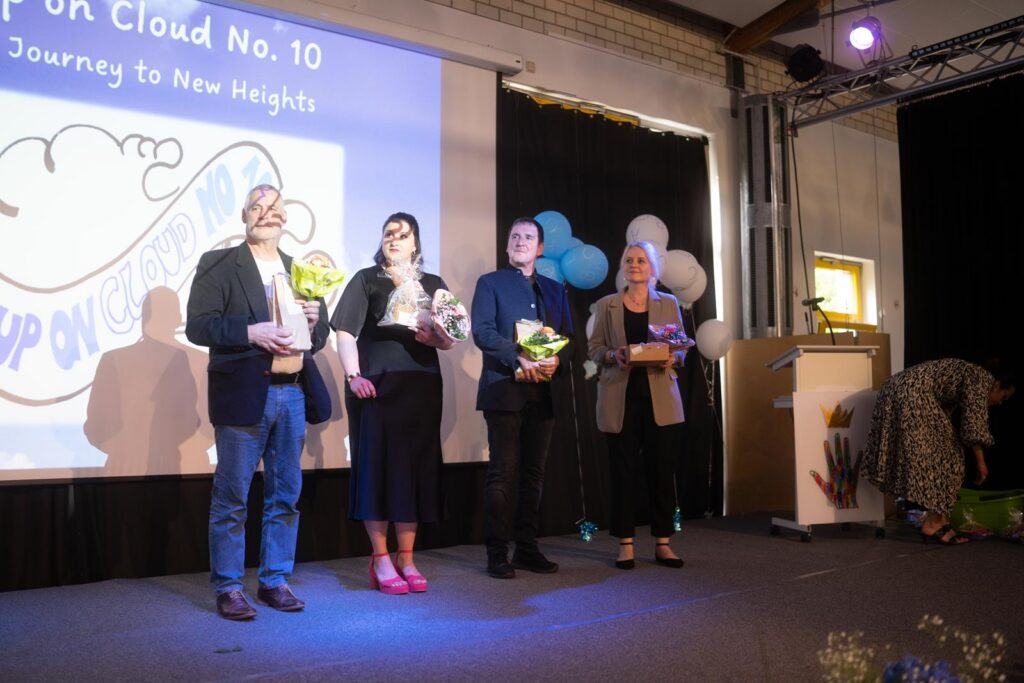 Vier Personen stehen auf einer Bühne mit Blumengeschenken. Hintergrund mit Dekoration.