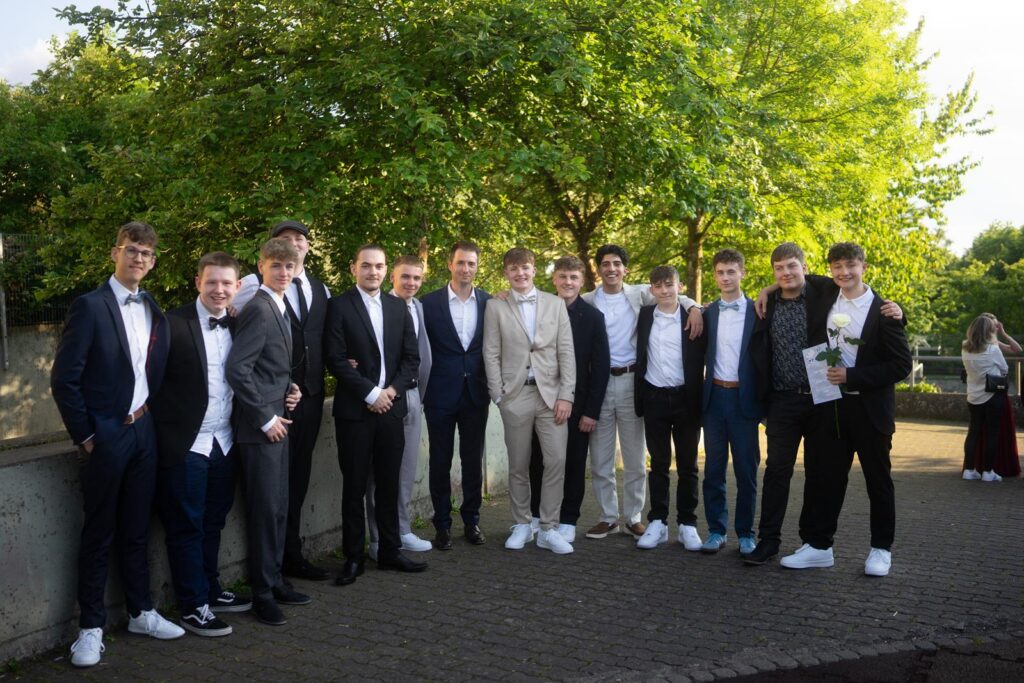 Gruppe von jungen Männern in Anzügen im Freien, mit Bäumen im Hintergrund.