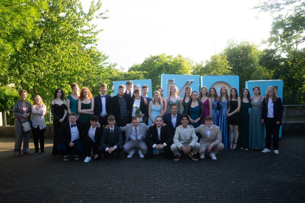 Gruppe von Schülern in festlicher Kleidung an einem Garten mit Bäumen.