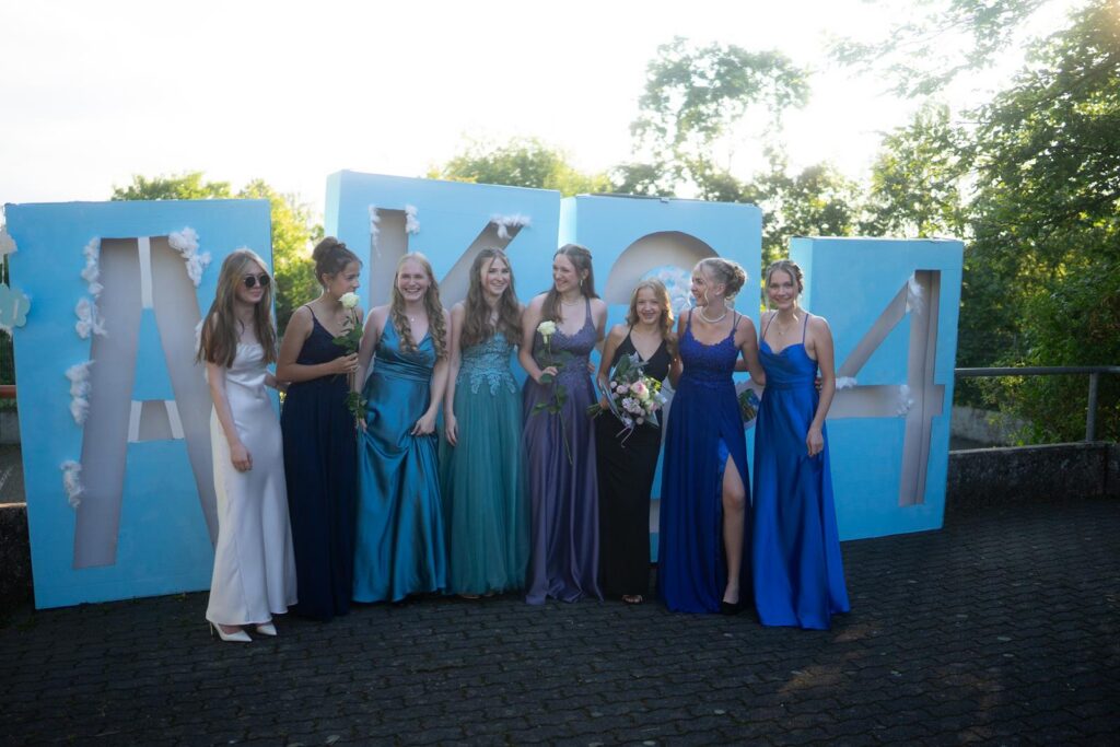 Gruppe von sieben Frauen in eleganten Kleidern vor großen bunten Zahlen.