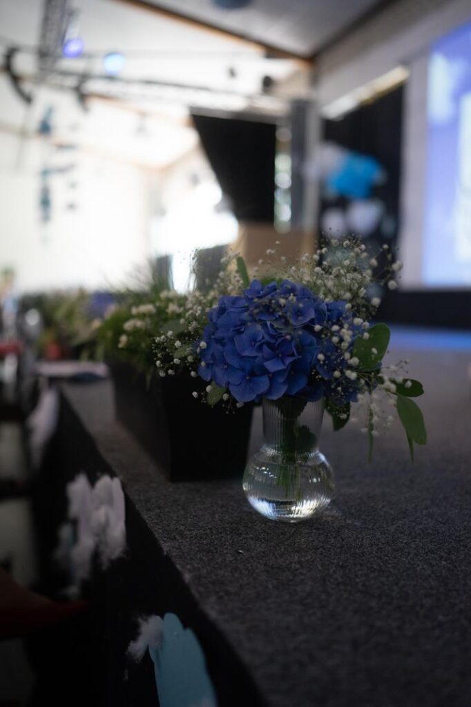 Blaue Blüten in einer Glasvase auf einem Tisch, im Hintergrund eine Bühne.