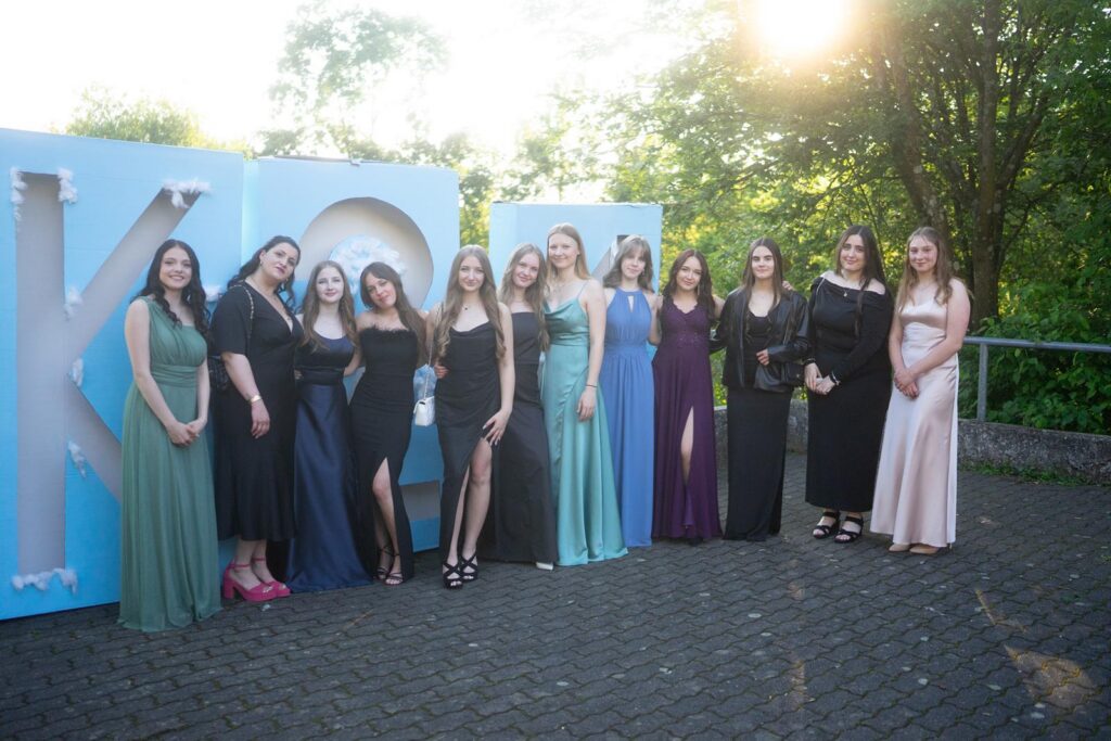 Gruppe von Frauen in festlicher Kleidung vor einem großen Schriftzug im Freien.