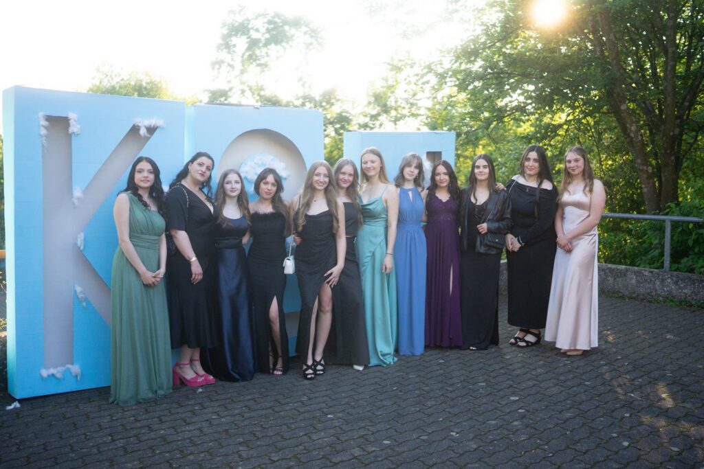 Gruppe von 13 Frauen in Abendkleidern vor einem großen Schriftzug "Koo".