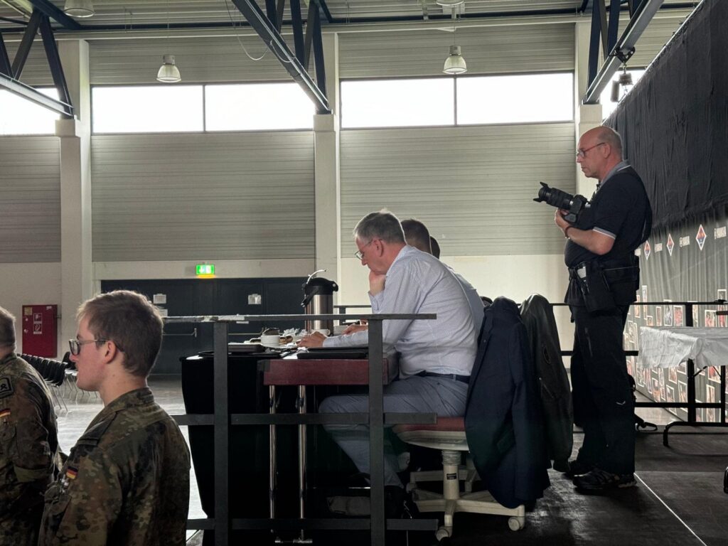 Personen sitzen an einem Tisch, im Hintergrund ein Sicherheitsbeamter mit Gewehr.