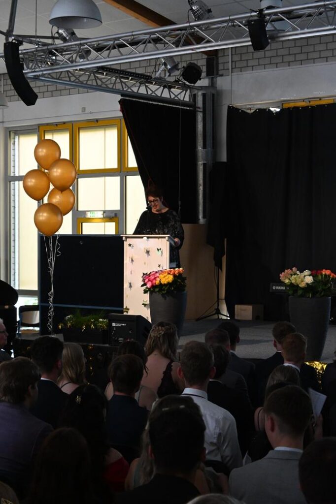 Person hält eine Rede auf einer Veranstaltung, umgeben von bunten Ballons und Blumen.
