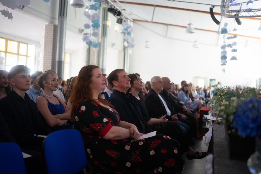 Zuschauer sitzen in einem Saal während einer Veranstaltung. Dekoration und Blumen sind sichtbar.