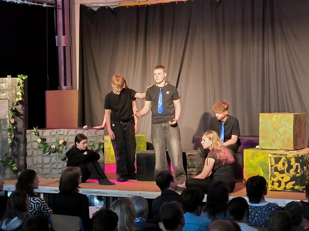 Schüler führen ein Theaterstück auf einer Bühne auf. Zuschauer sitzen davor.