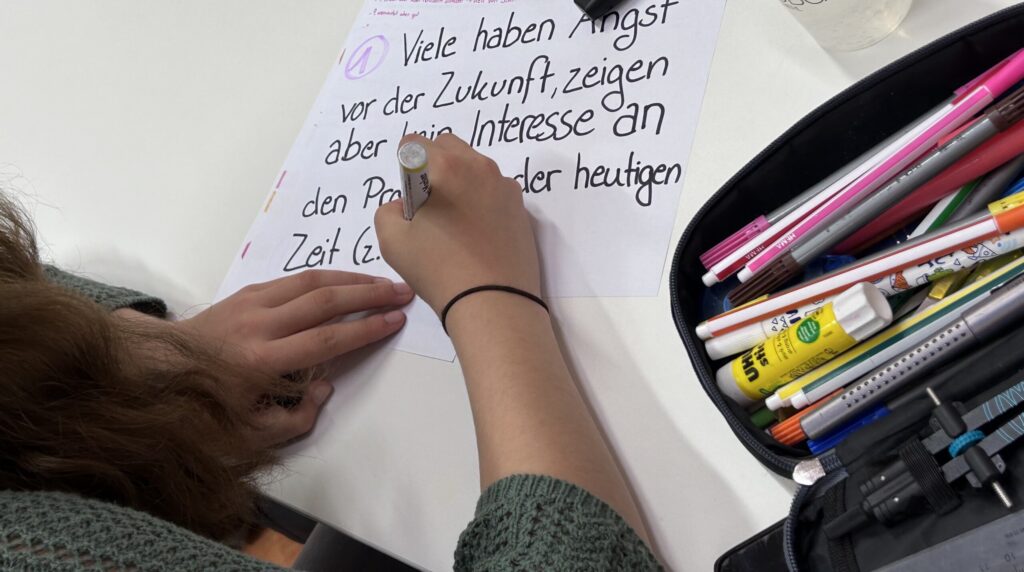 Hand schreibt auf Papier mit bunten Stiften neben einer Federtasche.