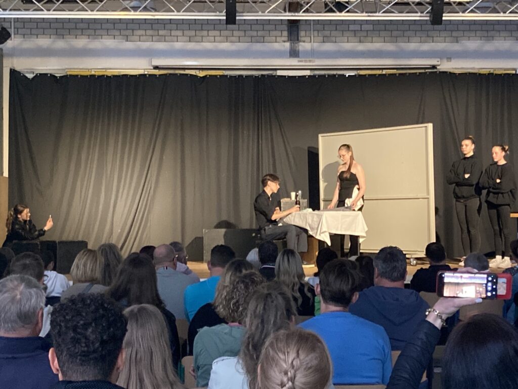 Theateraufführung mit mehreren Schauspielern auf der Bühne und Publikum im Vordergrund.