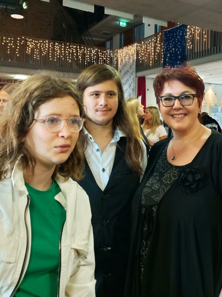 Drei Personen stehen in einer festlichen Umgebung und lächeln in die Kamera.