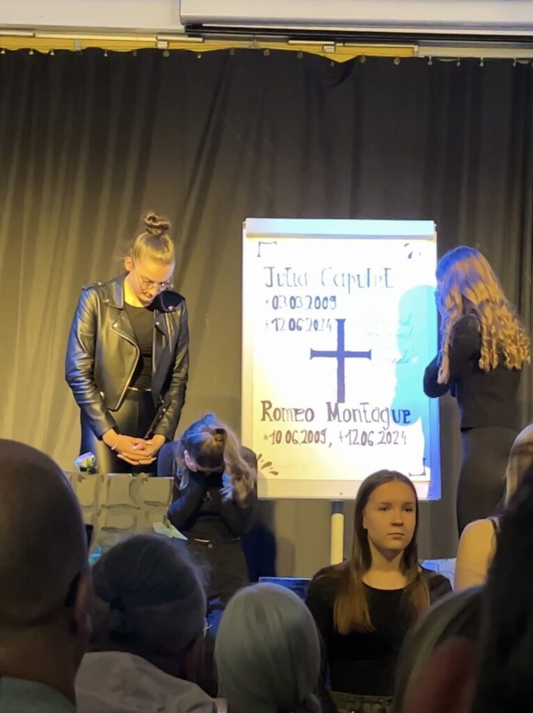 Trauerfeier mit zwei stehenden Personen und einem Gedenkbanner im Hintergrund.