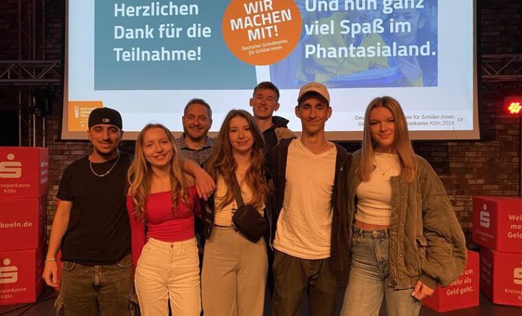 Gruppe junger Menschen posiert vor einer Präsentation auf einer Bühne.