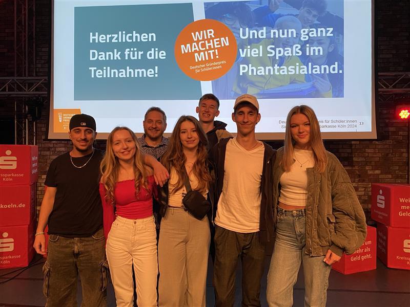 Gruppe junger Menschen posiert vor einer Präsentation auf einer Bühne.
