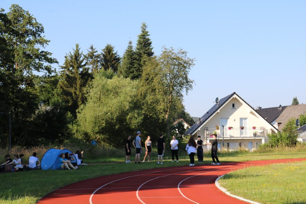 Gruppierungen von Personen am Sportplatz, im Hintergrund Bäume und Wohnhäuser.