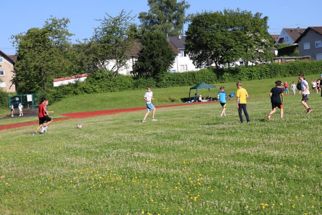 Schüler spielen auf einem Rasenfeld Fußball bei sonnigem Wetter.