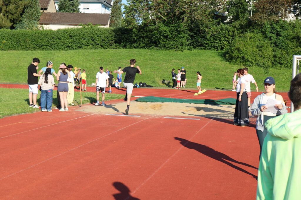 Schüler üben Leichtathletik auf dem Sportplatz. Sandgrube und Laufbahn sind sichtbar.