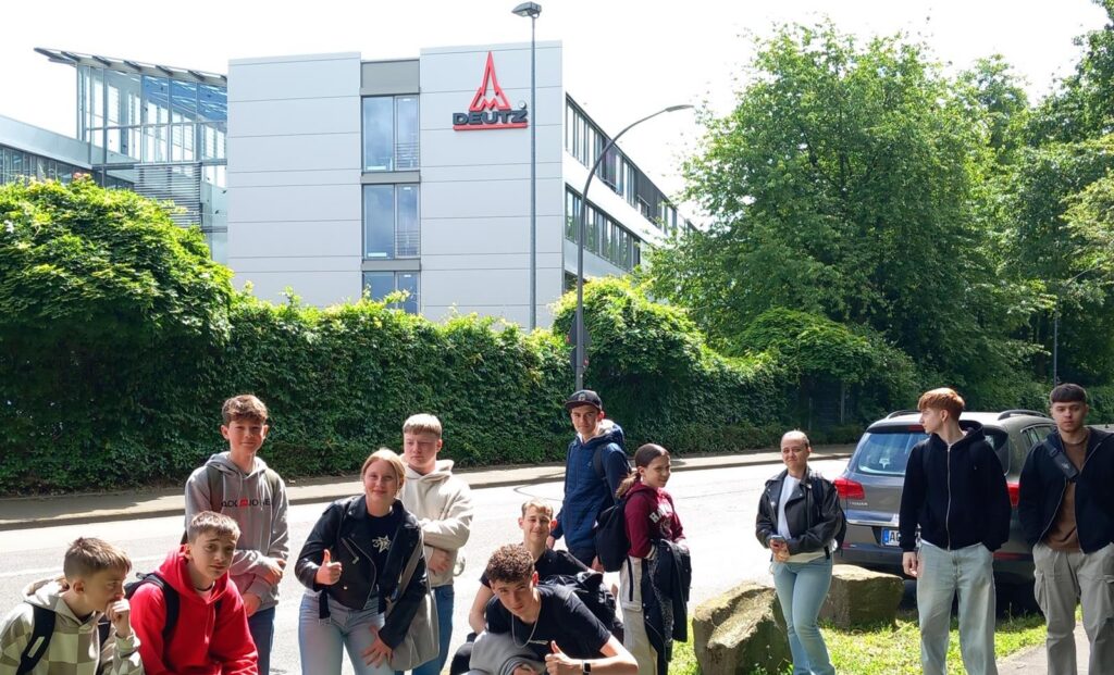 Gruppe von Schülern vor einem Gebäude mit Logo und Bäumen im Hintergrund.