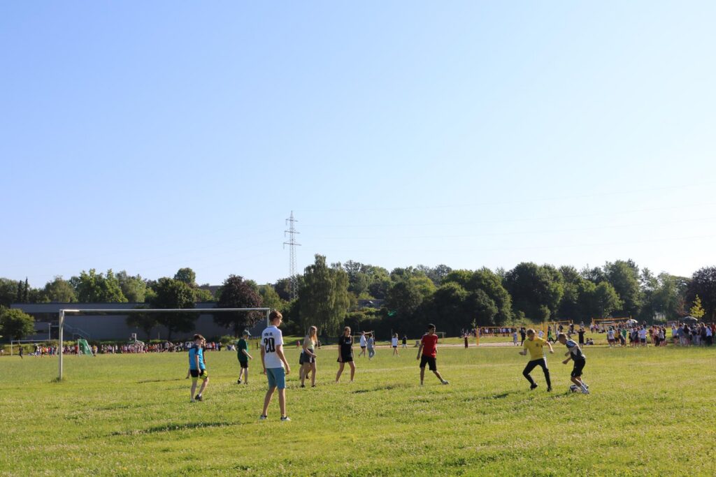 Kinder spielen Fußball auf einem grünen Feld unter blauem Himmel.