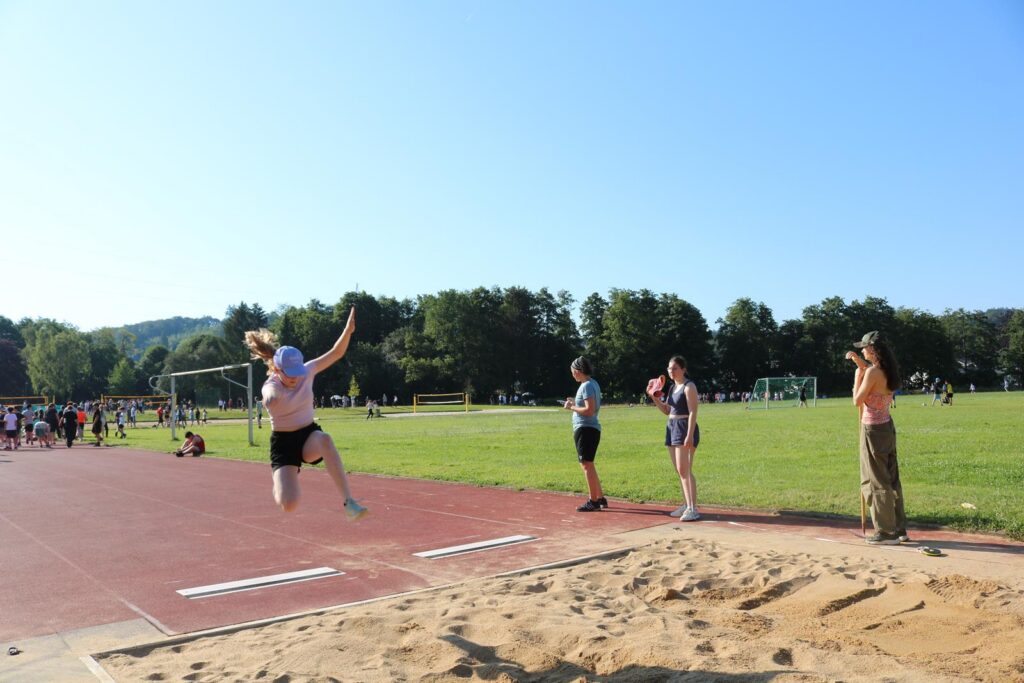 Sportler springt beim Weitsprung in den Sand. Zuschauer nehmen das Geschehen auf.