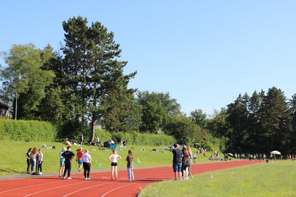 Sportplatz mit Laufbahn, Schüler und Schülergruppen am Rand, Bäume im Hintergrund.