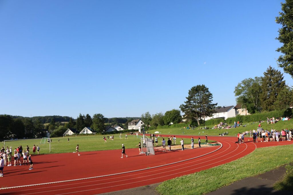 Sportplatz mit Laufbahn, Wiese und zahlreichen Menschen bei Aktivitäten.