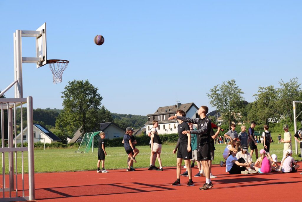 Schüler spielen Basketball auf einem Sportplatz bei sonnigem Wetter.