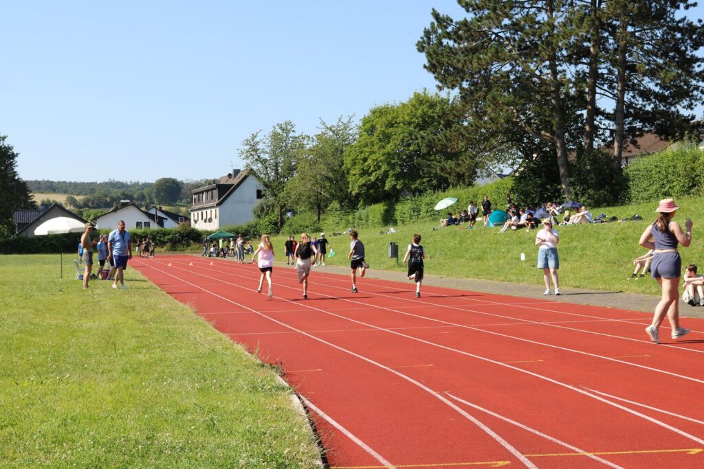 Schüler laufen auf einer roten Laufbahn, Zuschauer sitzen im Hintergrund auf der Wiese.