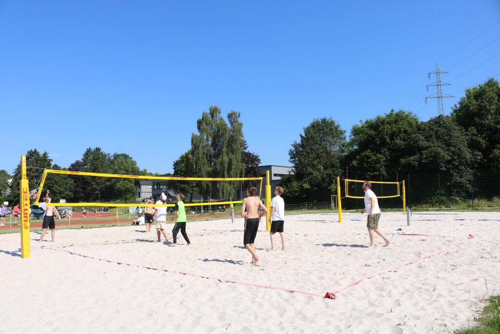 Personen spielen Volleyball auf einem Sandplatz an einem sonnigen Tag.