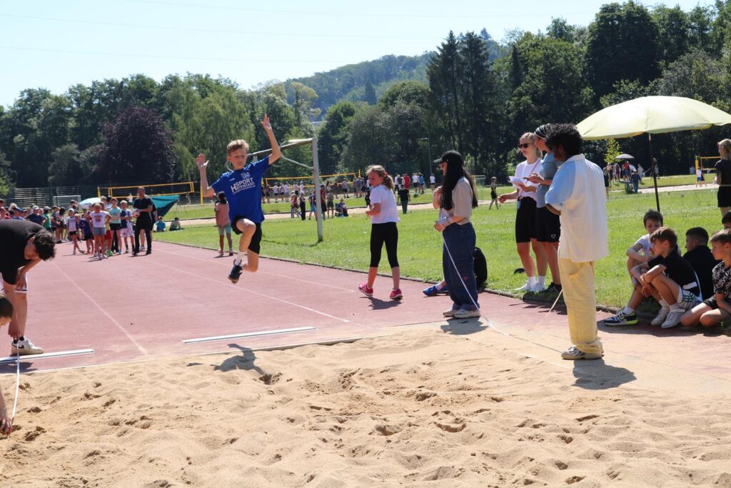 Junge springt in eine Sandgrube bei einem Sportevent, Zuschauer beobachten.
