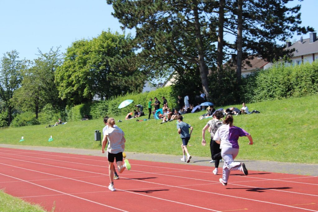 Schüler laufen auf einer Sportbahn, Zuschauer sitzen auf der Wiese.