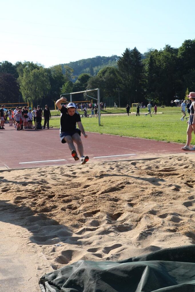 Ein Schüler springt in eine Sandgrube beim Sportevent. Zuschauer im Hintergrund.