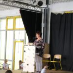 Eine Person steht am Mikrofon auf einer Bühne vor Publikum.