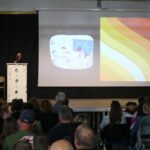 Vortragssituation mit Publikum, Präsentation im Hintergrund auf einer Bühne.
