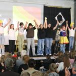Schülerinnen und Schüler stehen auf der Bühne und halten sich die Hände. Publikum applaudiert.