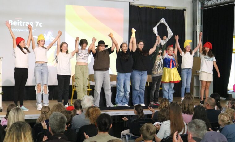 Schülerinnen und Schüler stehen auf der Bühne und halten sich die Hände. Publikum applaudiert.