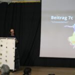 Schülerin spricht an einem Rednerpult, Präsentation im Hintergrund.