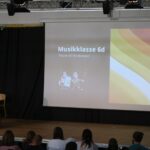Lehrer präsentiert in der Aula, im Hintergrund Folie der Musikklasse 6d.