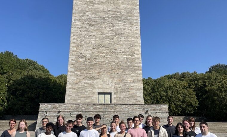 Gruppe von Schülern vor einem hohen, steinernen Denkmal bei klarem Himmel.