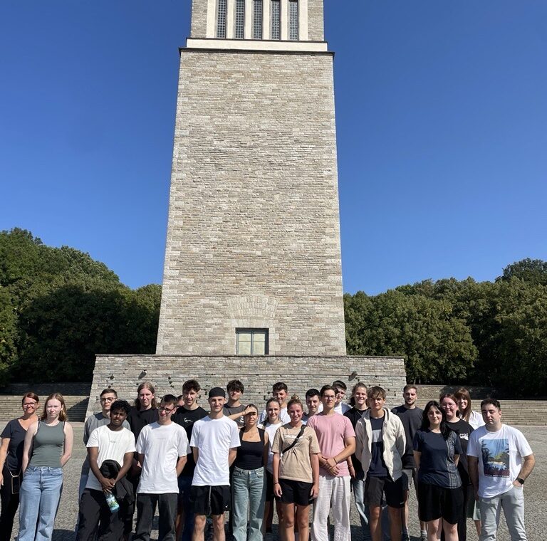 Gruppe von Schülern vor einem hohen, steinernen Denkmal bei klarem Himmel.