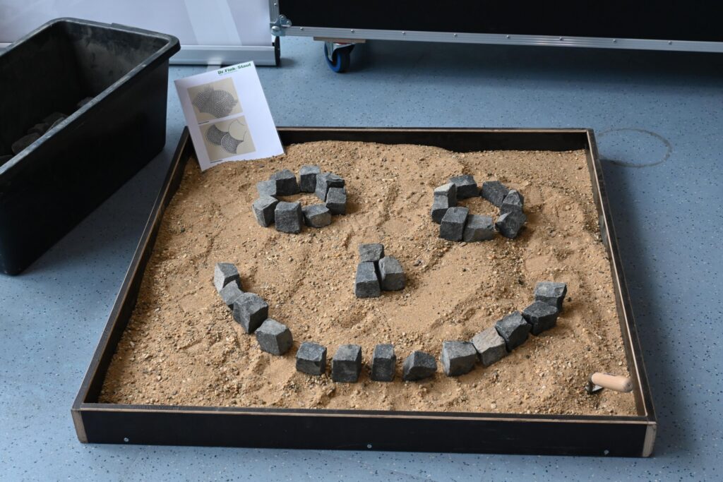Sandkasten mit Sand und Steinen, die ein Smiley-Gesicht bilden.