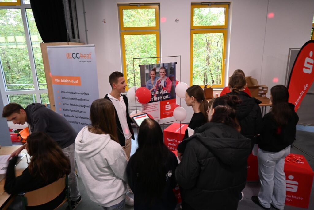 Messestand mit Schülern, Informationsmaterial und Dekoration, im Hintergrund ein Plakat.
