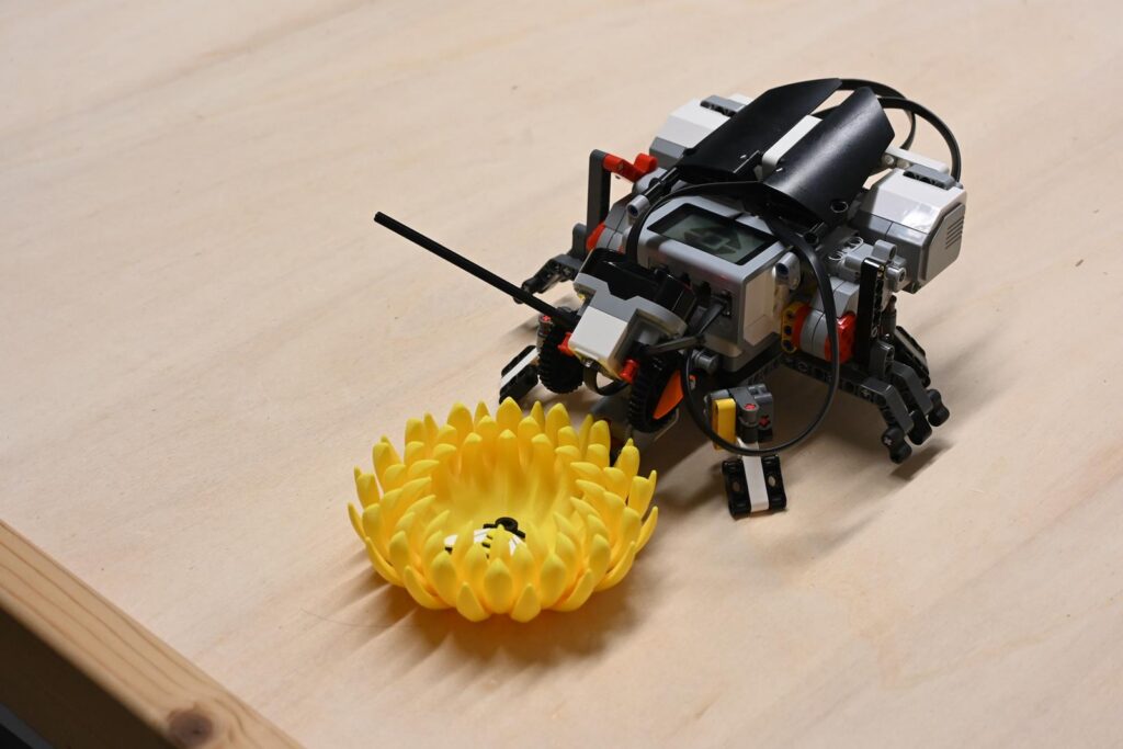 Ein Roboter steht neben einem gelben, blumenförmigen Objekt auf einem Tisch.