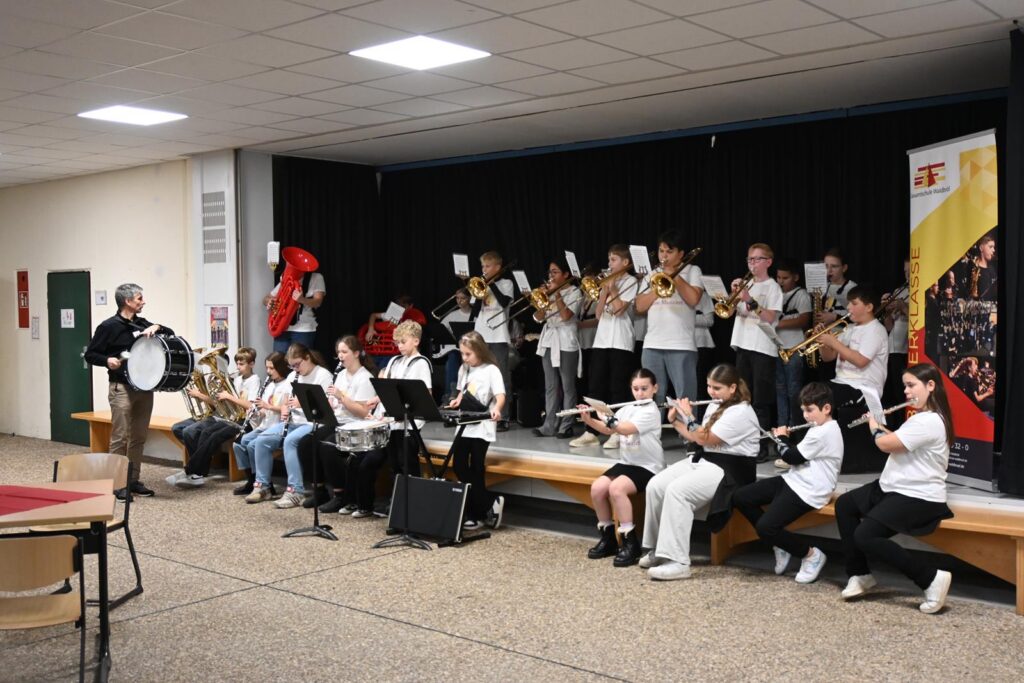 Schüler in weißen T-Shirts spielen Musikinstrumente auf einer Bühne.