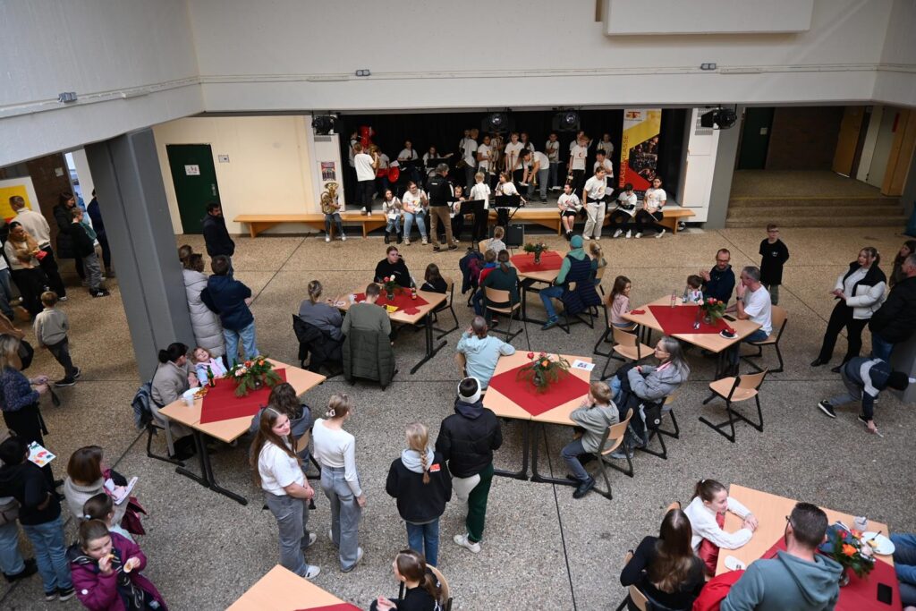 Schüler und Besucher sitzen an Tischen in einer Schulveranstaltung, Bühne im Hintergrund.