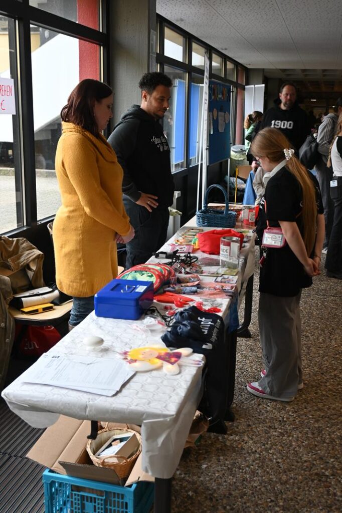 Stand mit Ausstellern und Besuchern in einem Flur. Verschiedene Materialien sind auf einem Tisch.