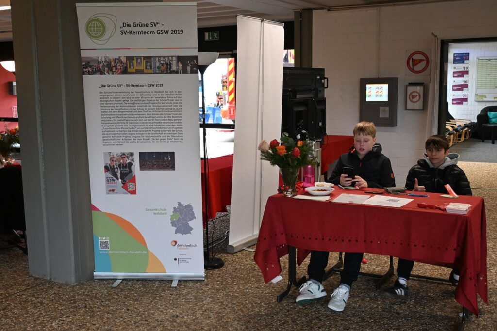 Zwei Schüler sitzen an einem Tisch mit einer Informationswand im Hintergrund.